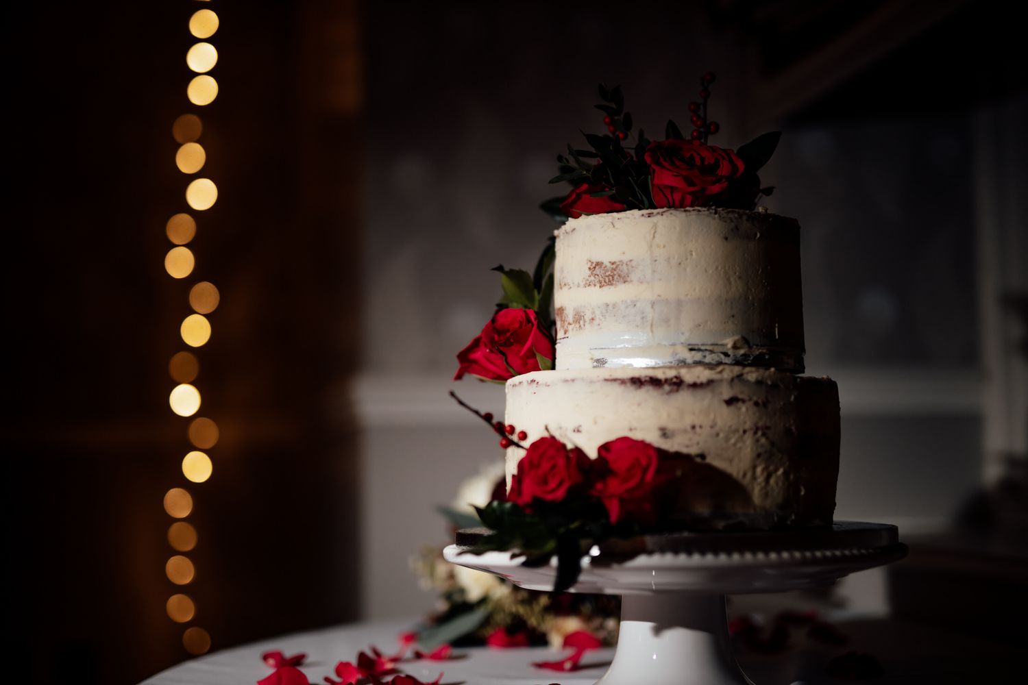 White wedding cake decorated with red roses against a dark background with twinkle lights.