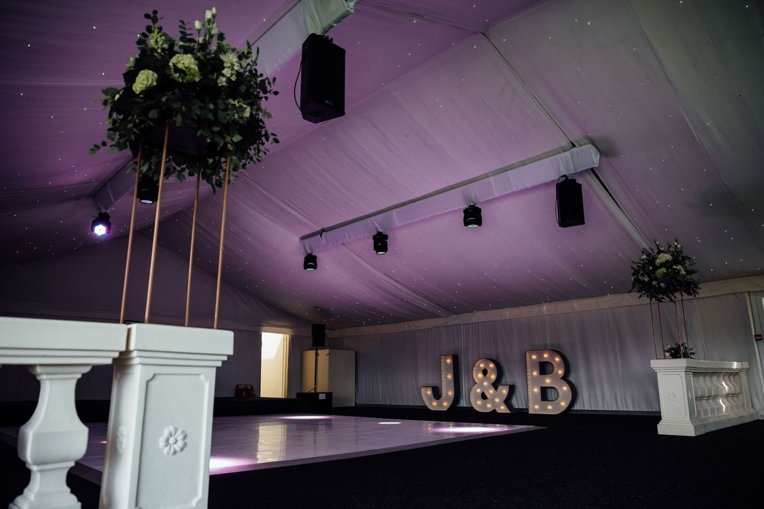 Wedding reception venue with purple lighting, floral arrangements and 'J&B' letters displayed on the wall.