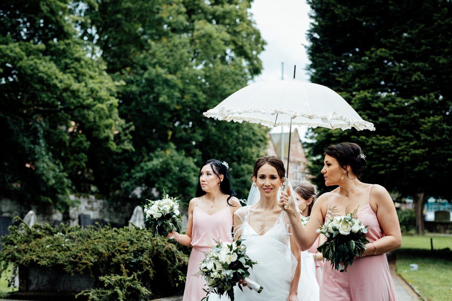 Wedding party in pink bridesmaid dresses poses together outdoors with parasols and bouquets.