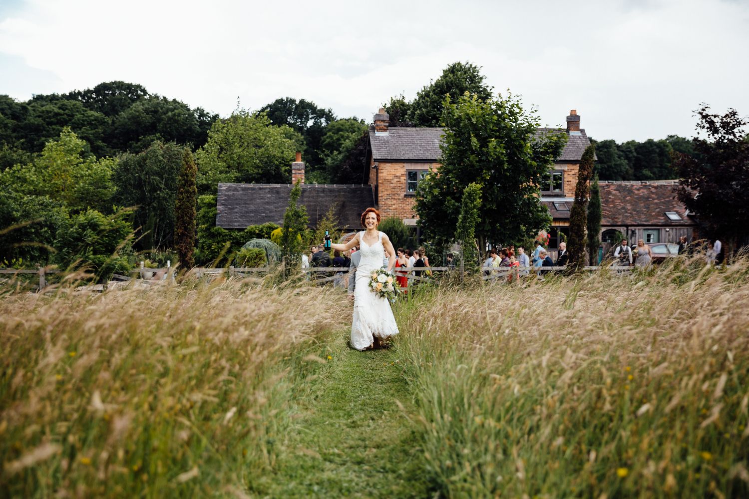 A bride walking through tall grass with rustic brick buildings in the background.
