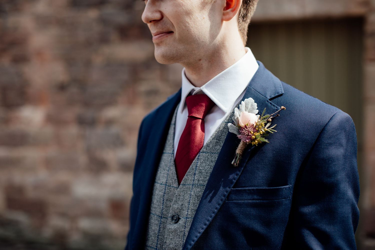 Groom in navy suit with red tie stands outside brick venue.