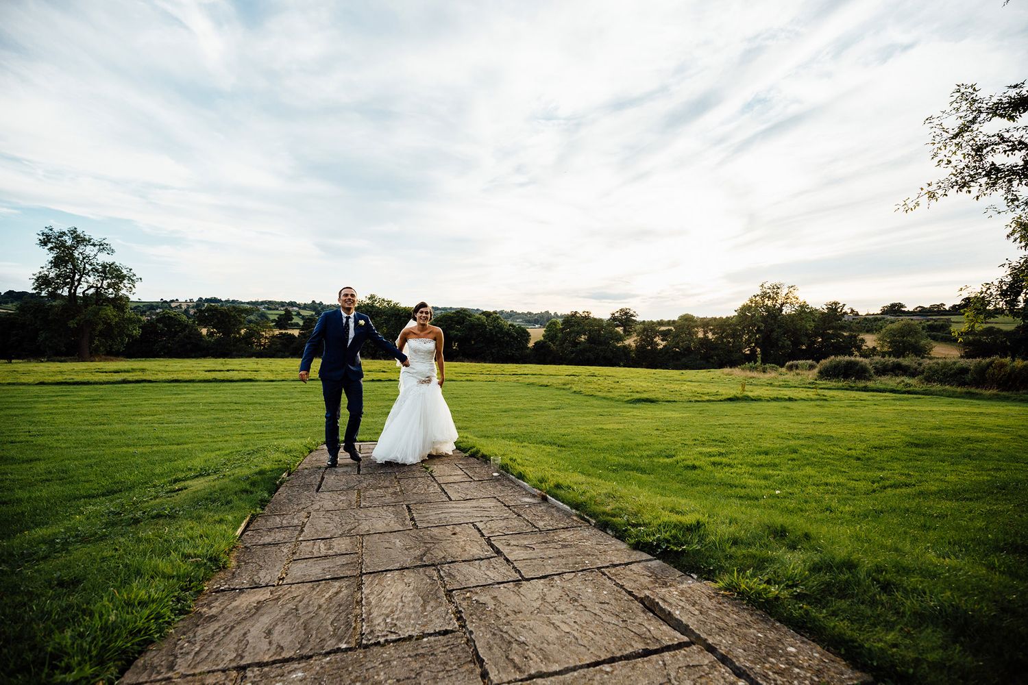 Couple walks along stone path through green meadow during sunset wedding photos.