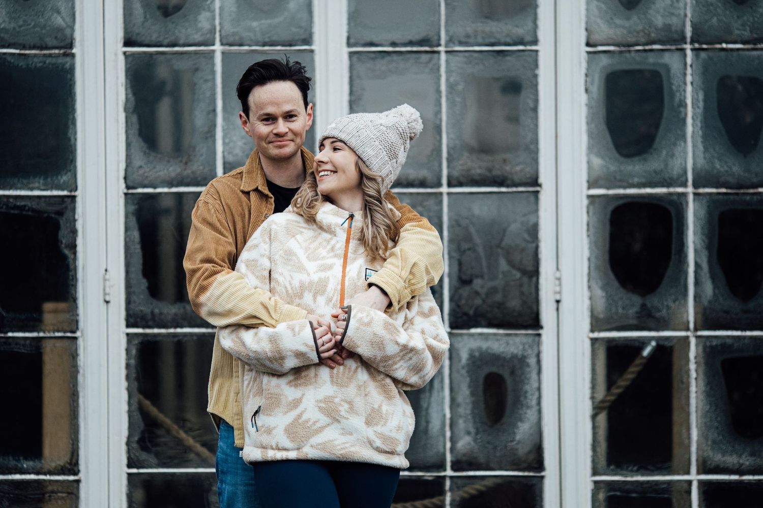 A couple embracing and laughing together in front of a glass window wall.