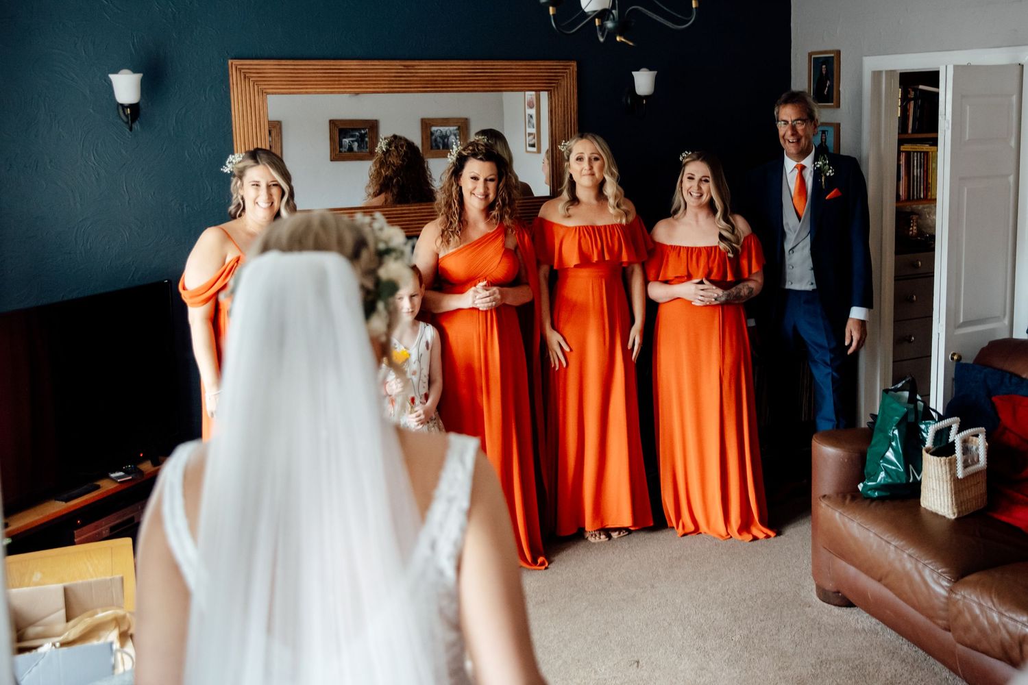 Bridesmaids in matching orange dresses react emotionally to seeing the bride in her gown.