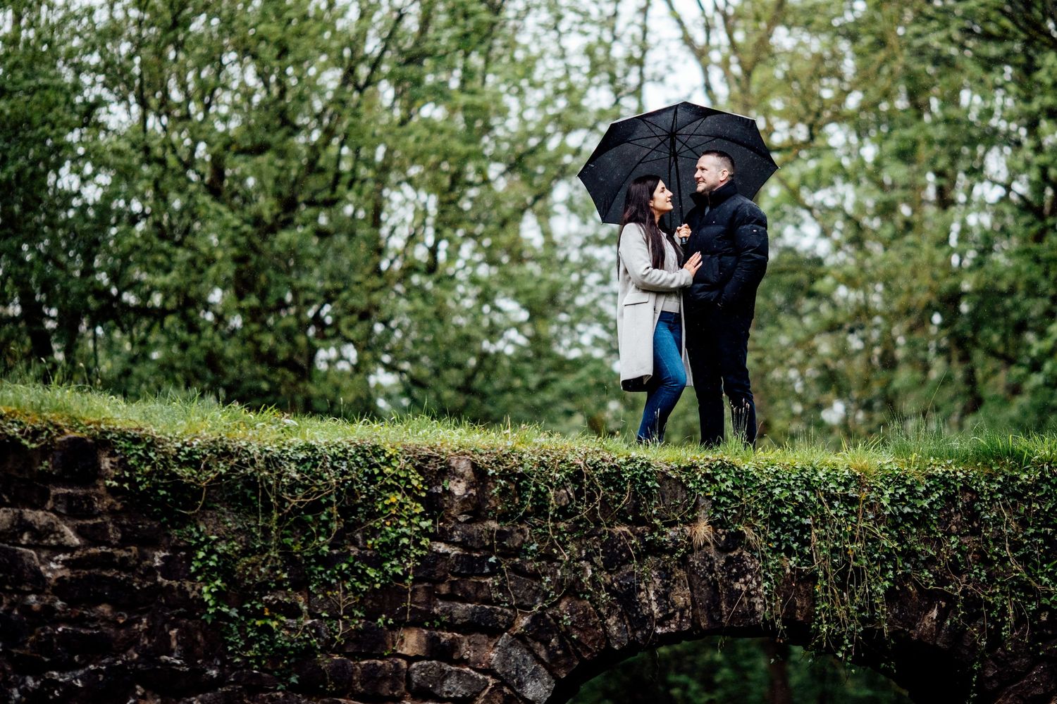 A couple stands together under an umbrella on a mossy stone wall in the woods.