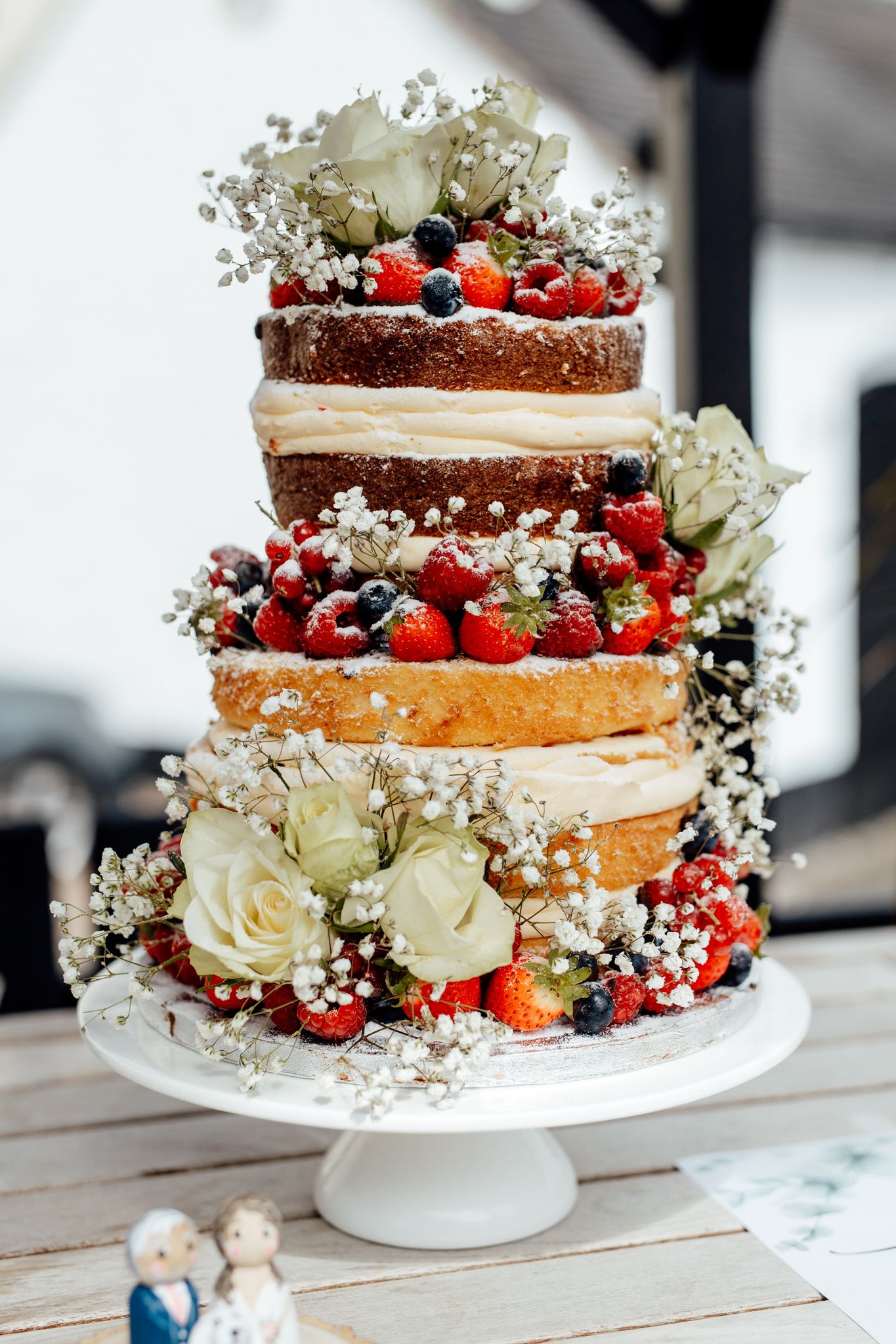 A rustic naked wedding cake decorated with fresh berries, white roses and baby's breath on a white cake stand.