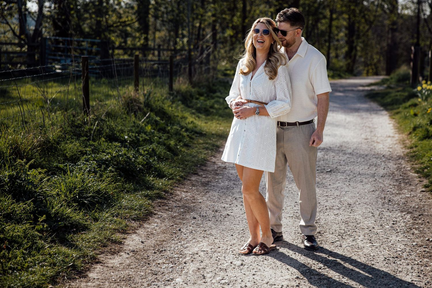 A romantic couple walks together on a gravel path through a scenic countryside setting.