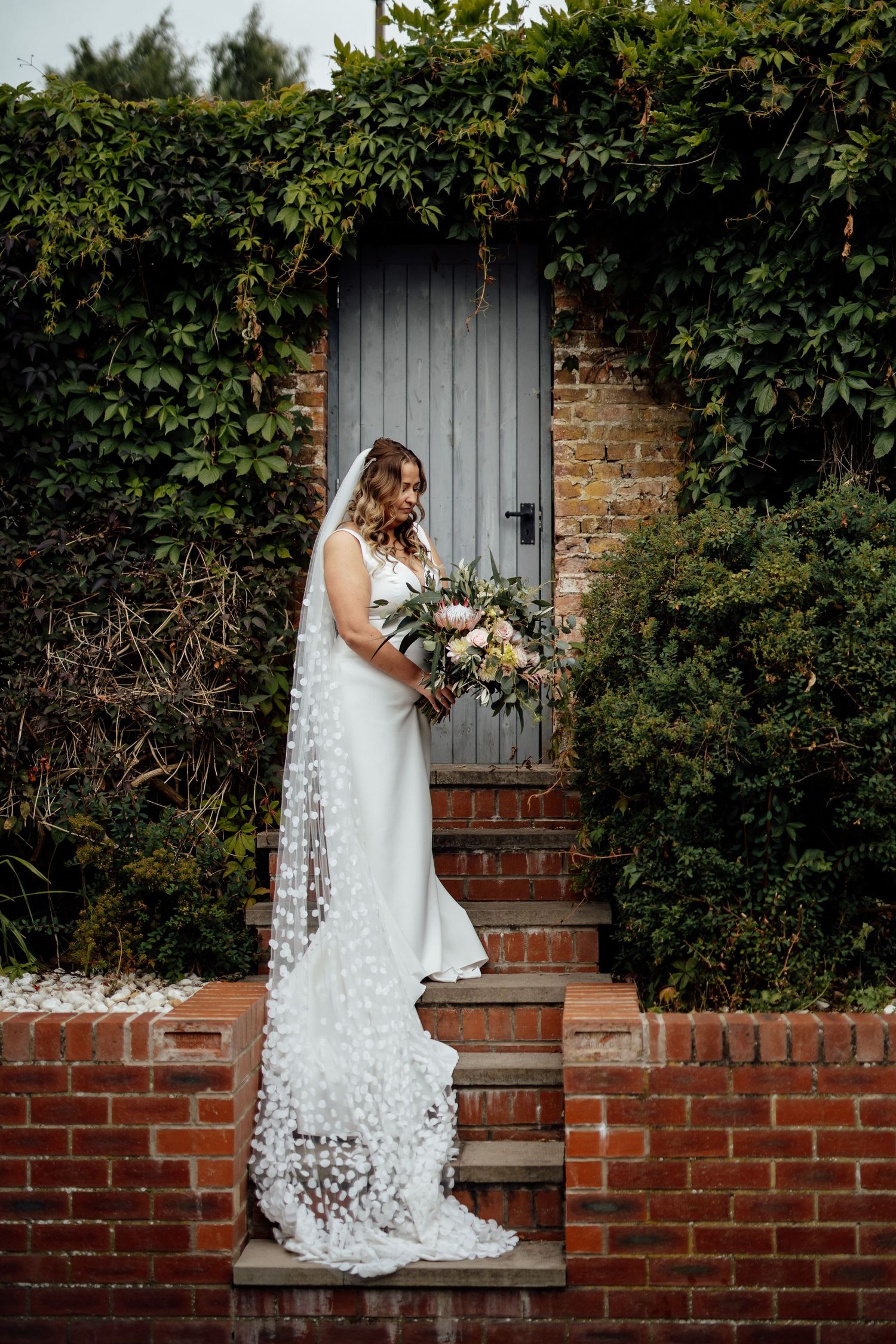 Elegant wedding dress with long lace train against rustic brick wall and wooden door covered in climbing vines.