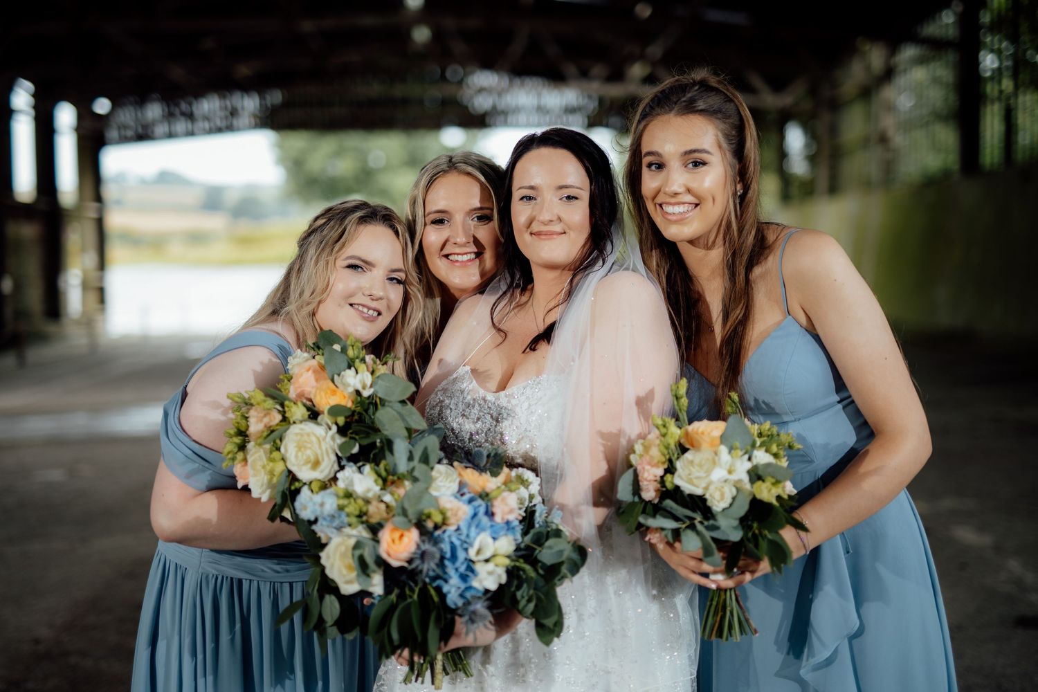 Bridesmaids in dusty blue dresses hold yellow and white bouquets while posing under a covered walkway.