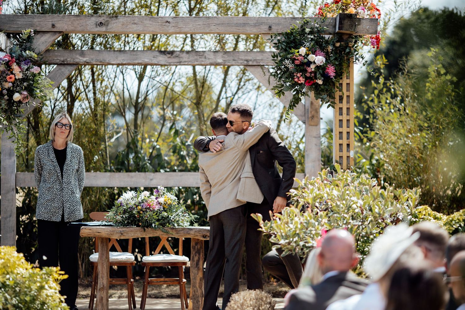 Two grooms embrace at an outdoor wedding ceremony surrounded by colorful floral arrangements.