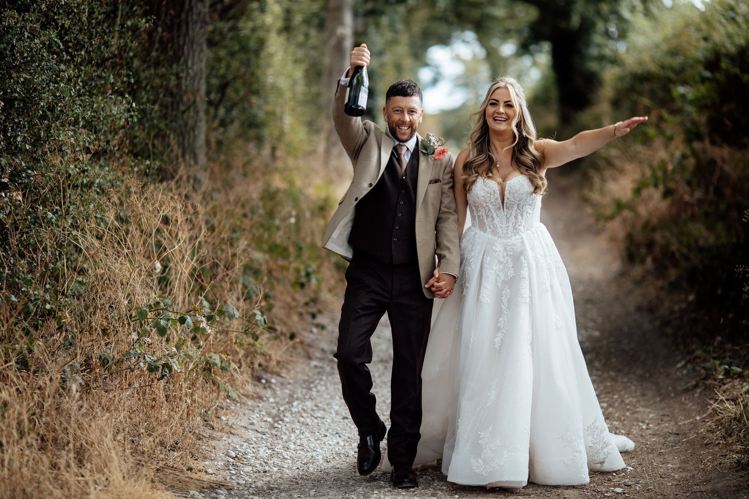 Happy newlyweds walk down a country lane as groom celebrates with champagne bottle.