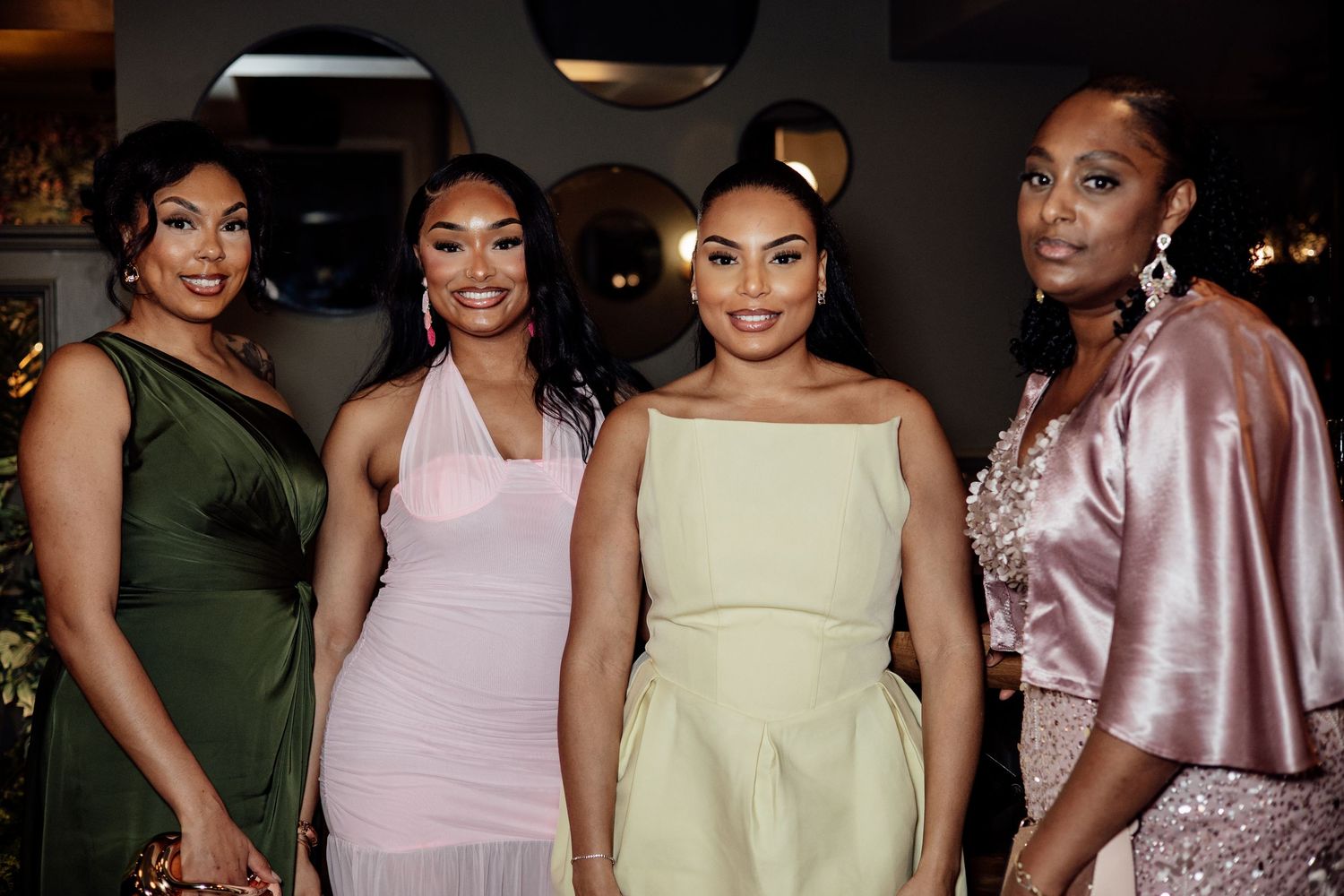 Four women in elegant formal dresses posing together at a wedding reception event.