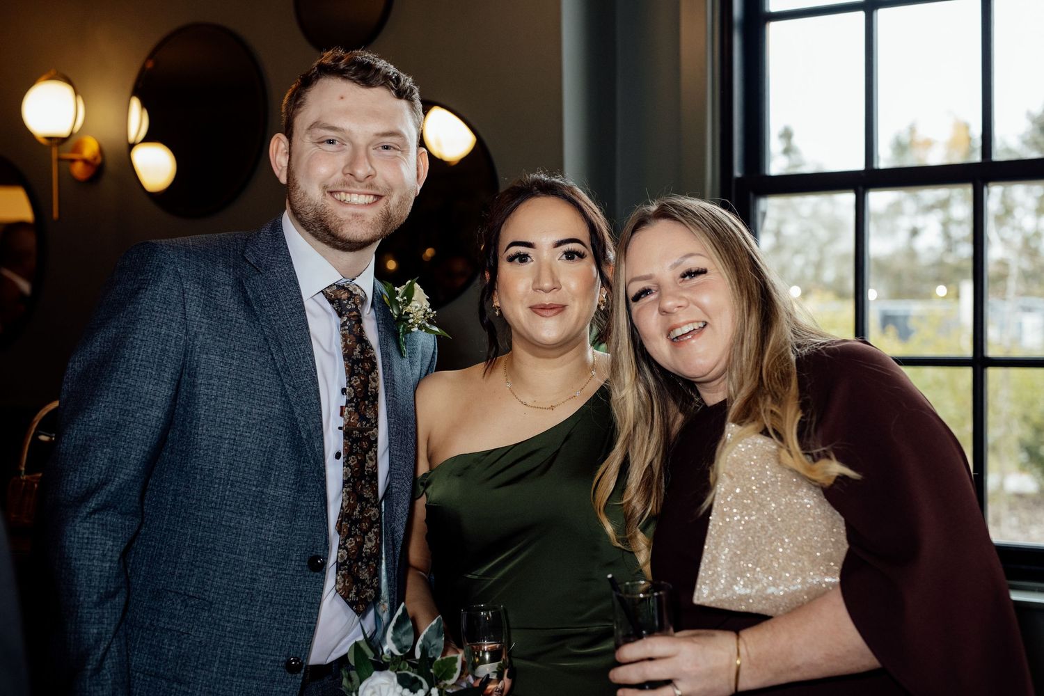 Three wedding guests smile together near a window at a wedding reception.