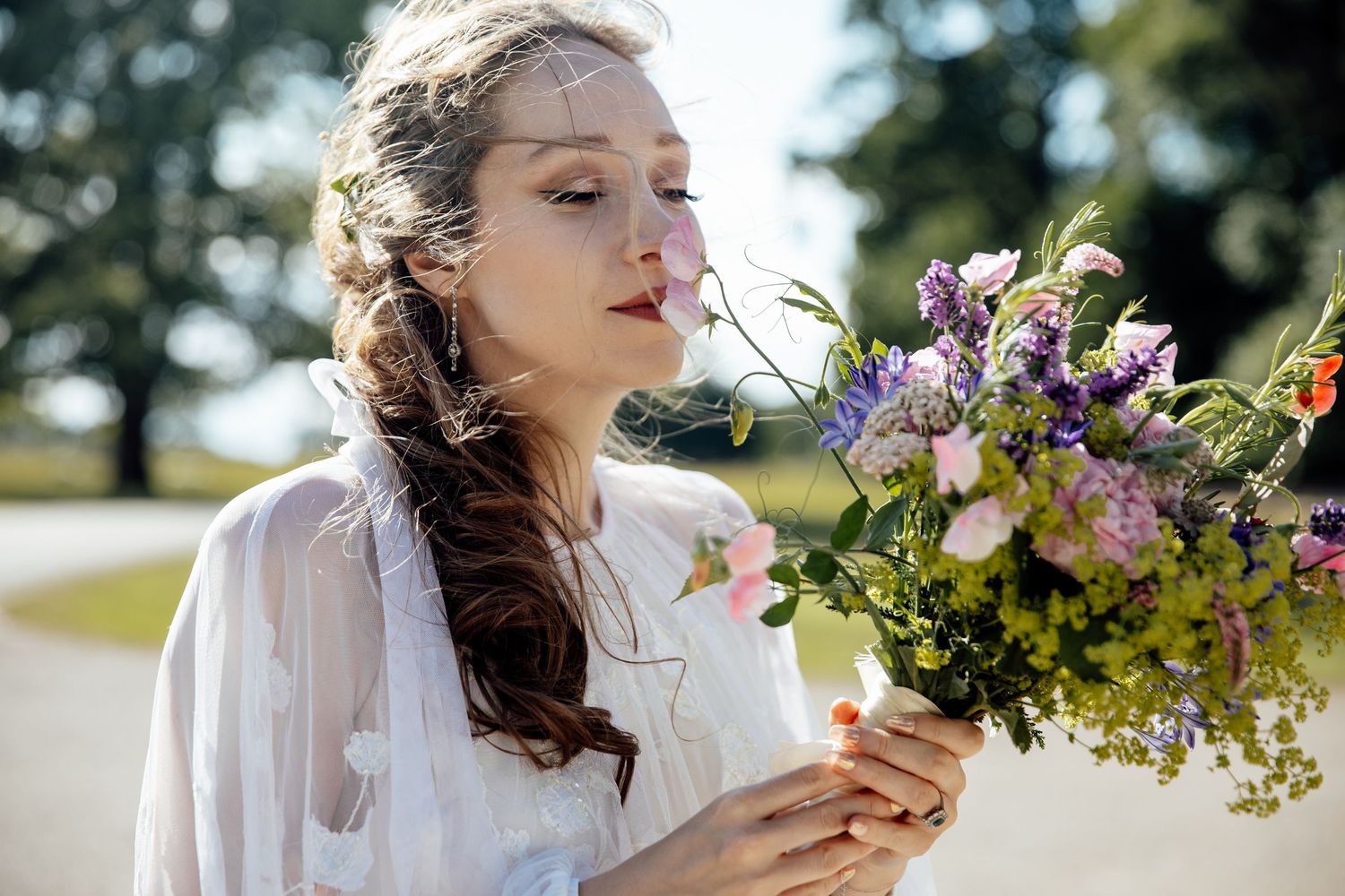 A woman in a white dress holds and smells a beautiful bouquet of wildflowers in sunlit outdoor setting.