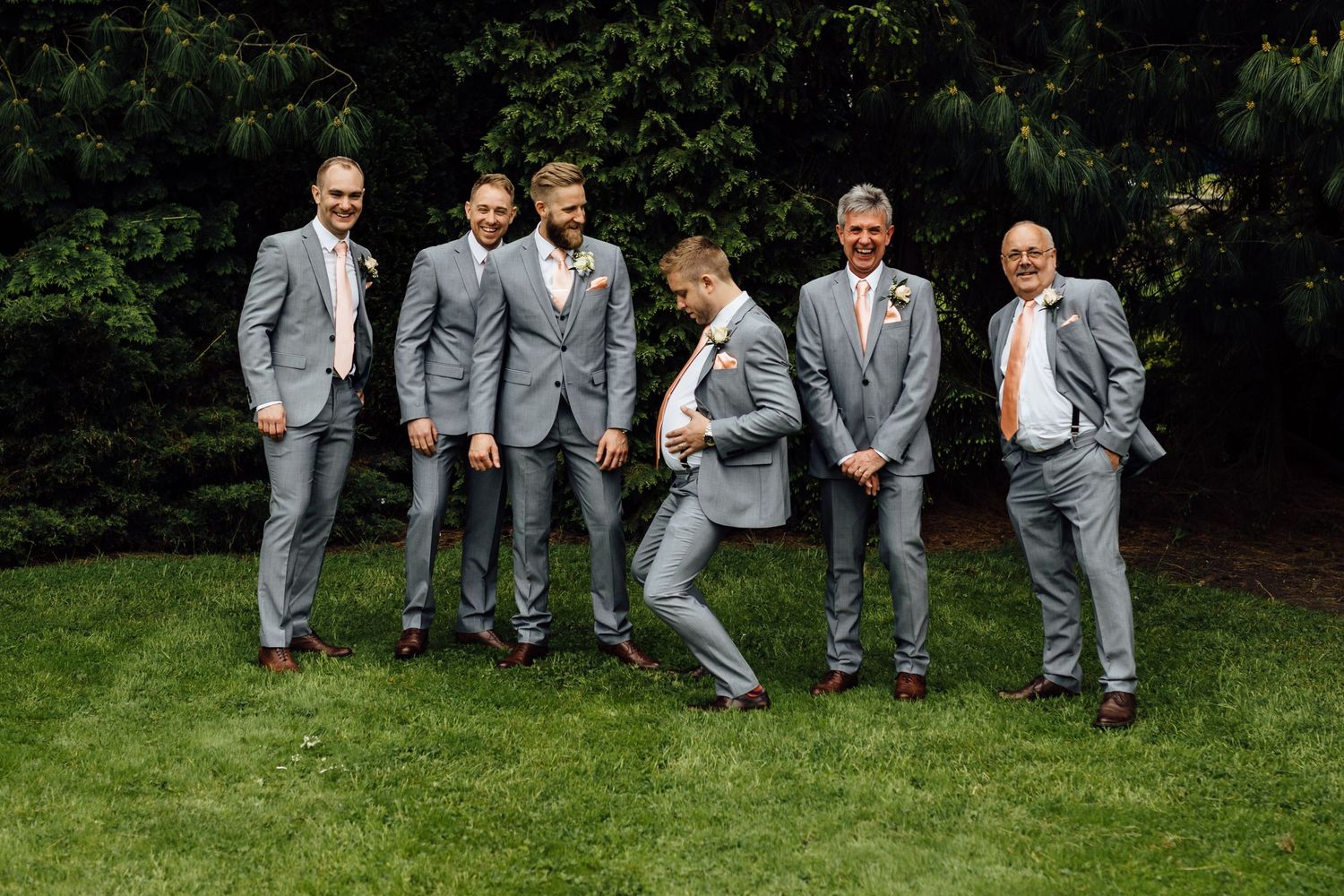 Groomsmen in gray suits pose playfully on green lawn for wedding photos.