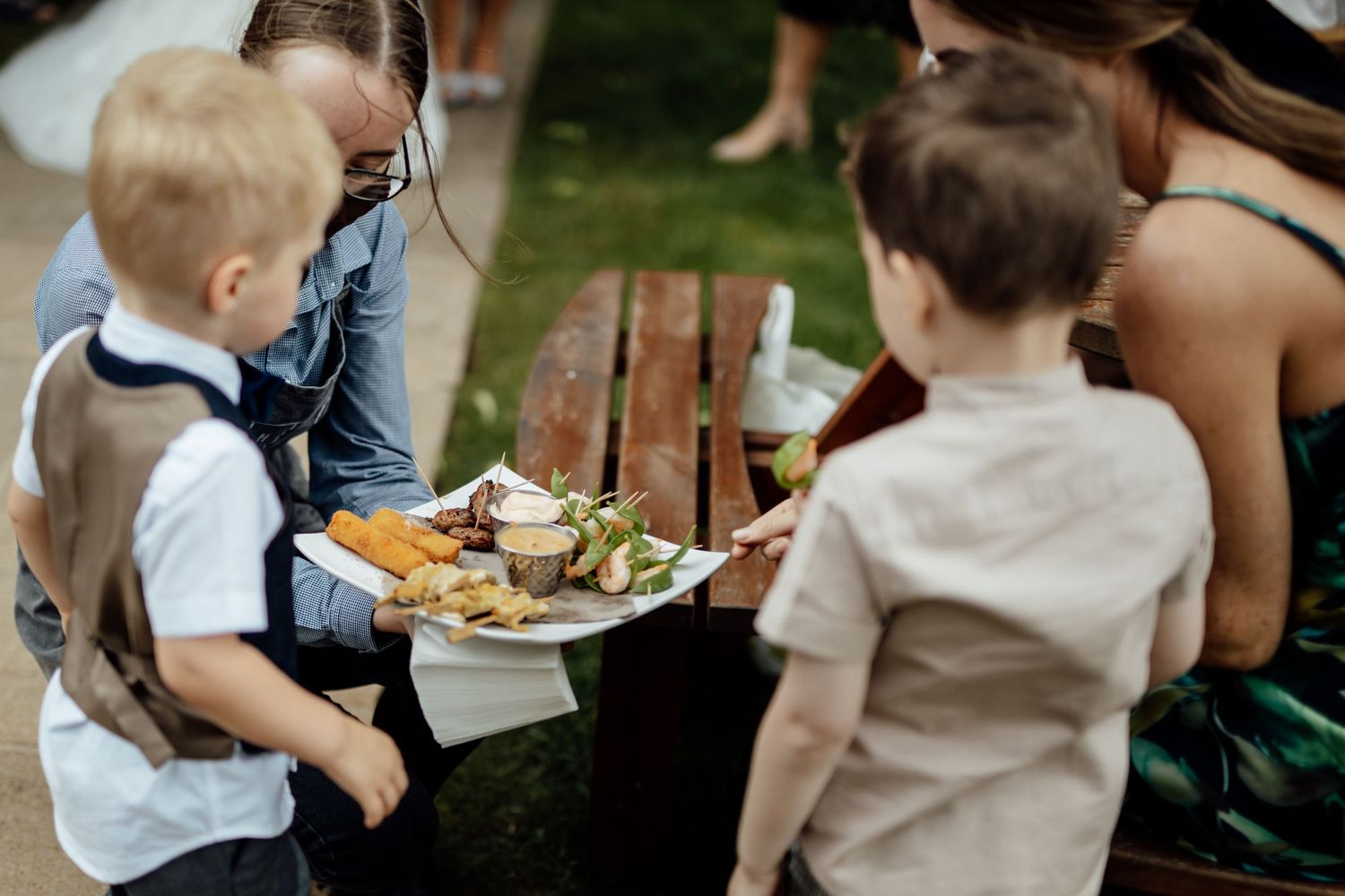 Children in formal attire gather around a buffet table at an outdoor wedding reception.