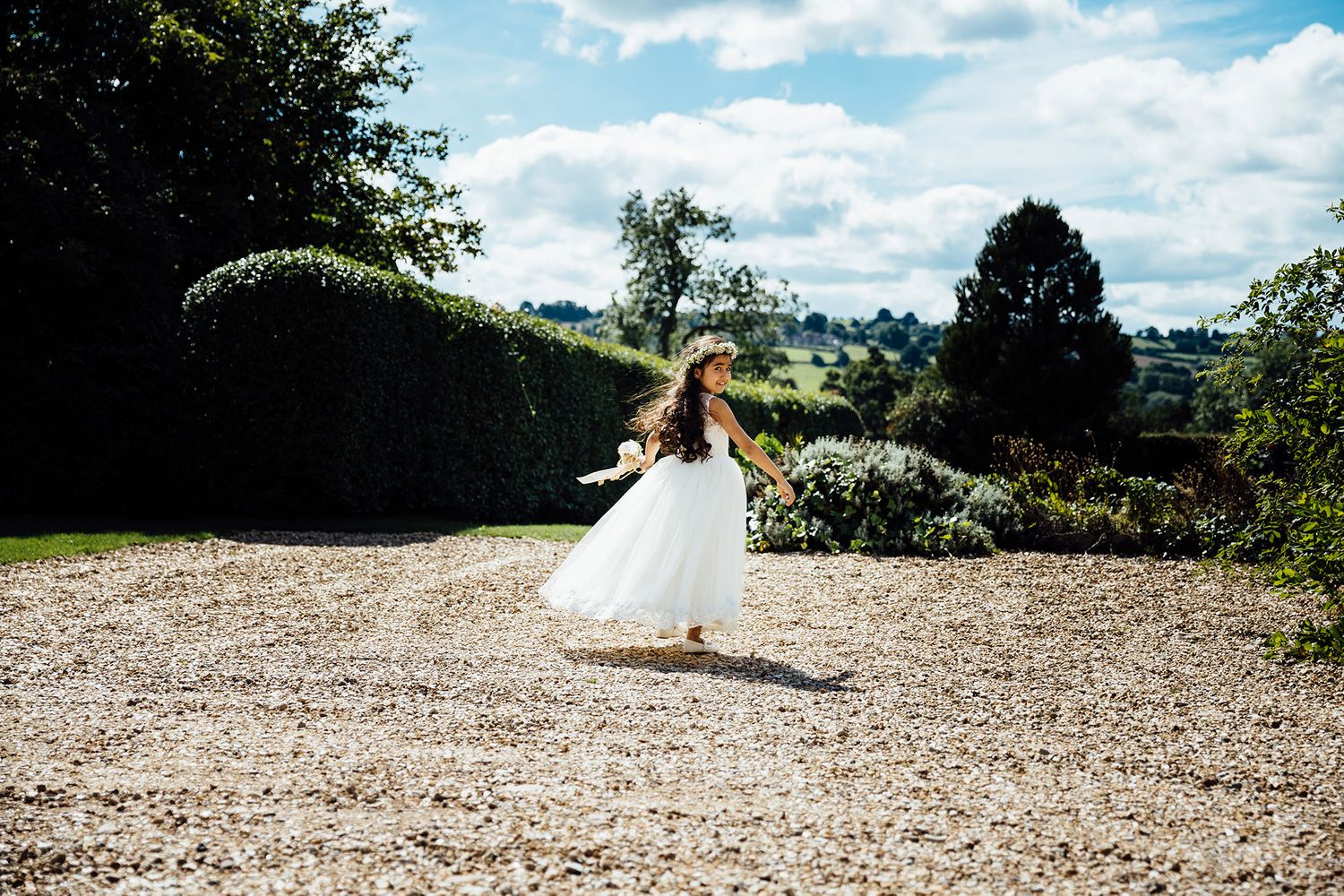 A person in a white dress twirls on a gravel path surrounded by garden scenery.