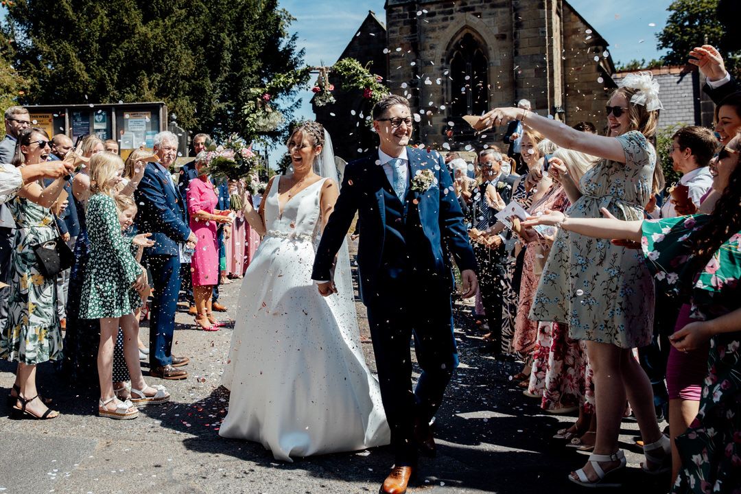 Wide angle shot of wedding guests creating a confetti tunnel outside a historic stone church.