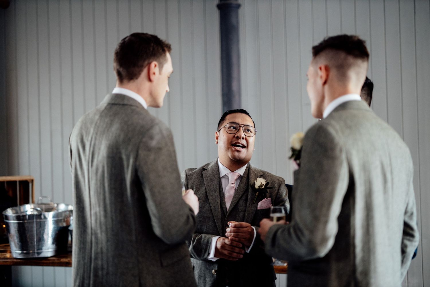 Three groomsmen in grey suits share a laugh during wedding preparations.