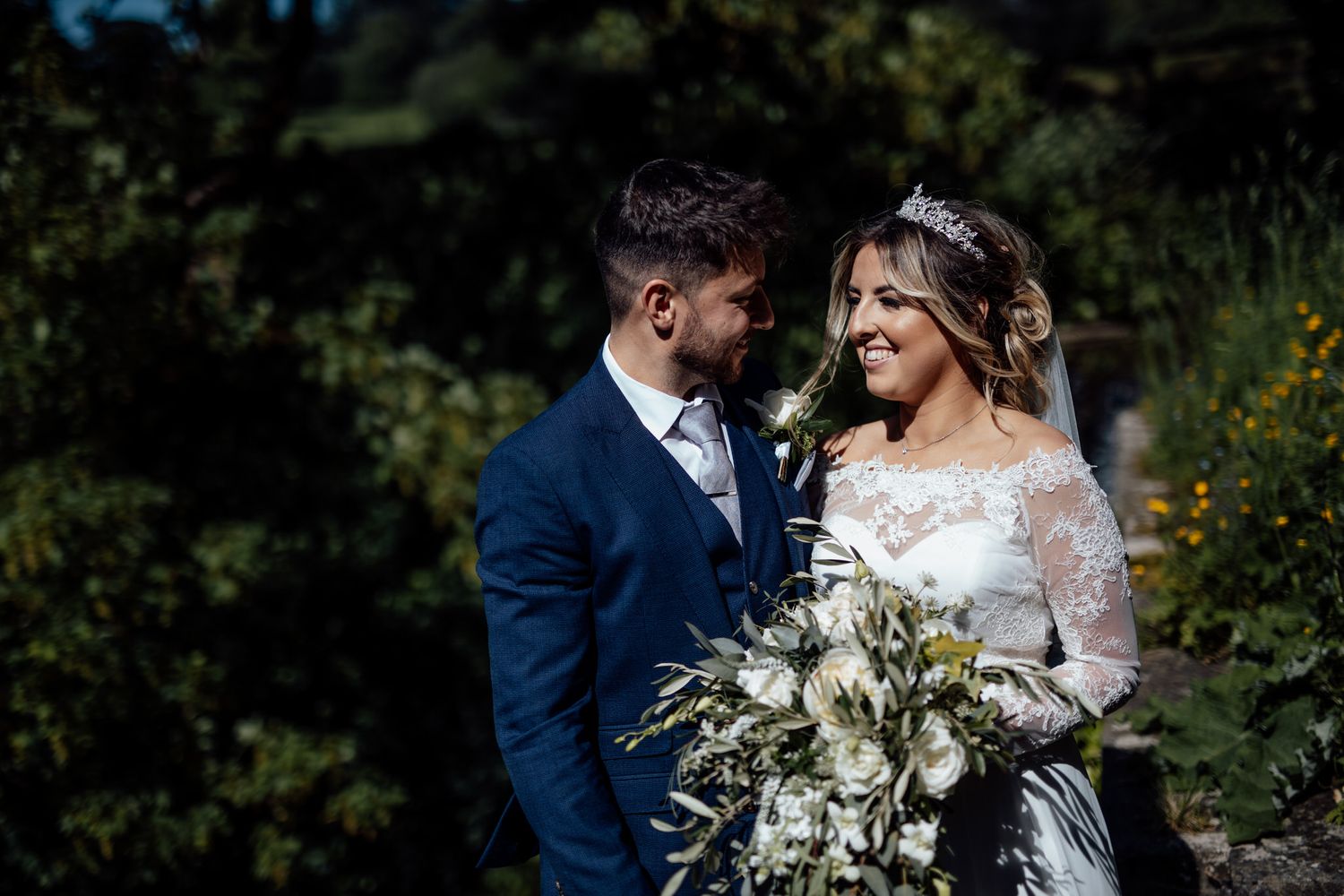 Elegant wedding couple posing with white floral bouquet in a garden setting.