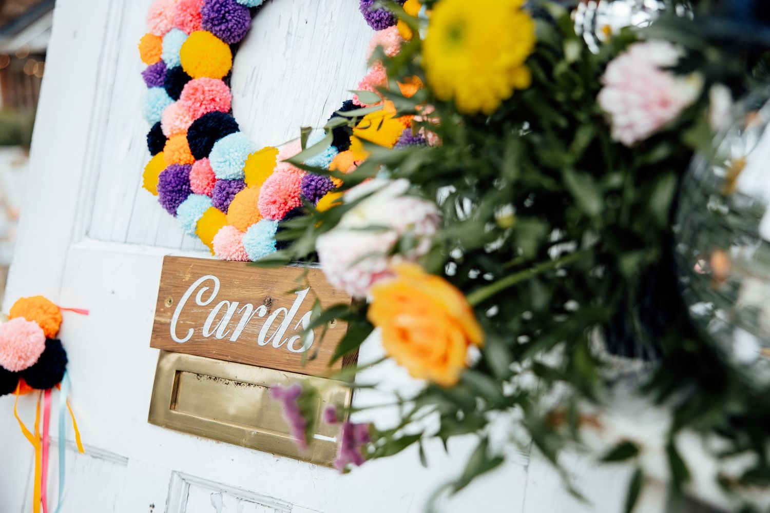 Decorative card box with colorful pom pom wreath and floral arrangements at wedding reception.