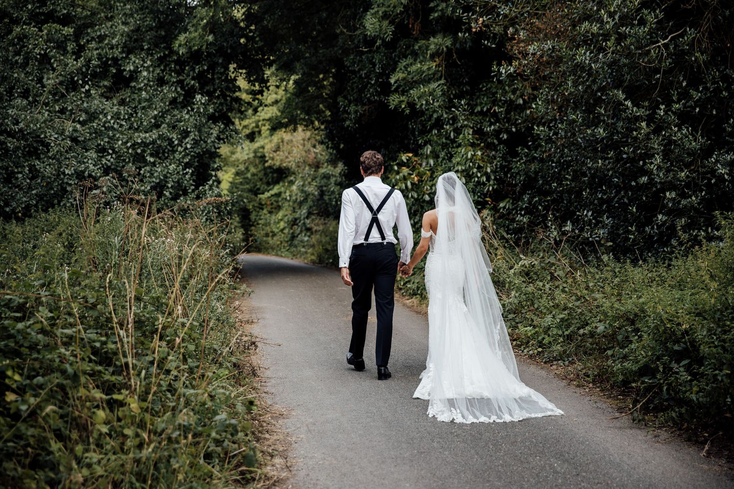 Couple walking down garden path in wedding attire with lace dress and suspenders.