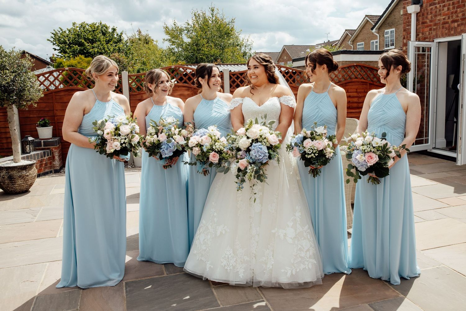 Bridal party in powder blue dresses poses outdoors with floral bouquets against brick building.