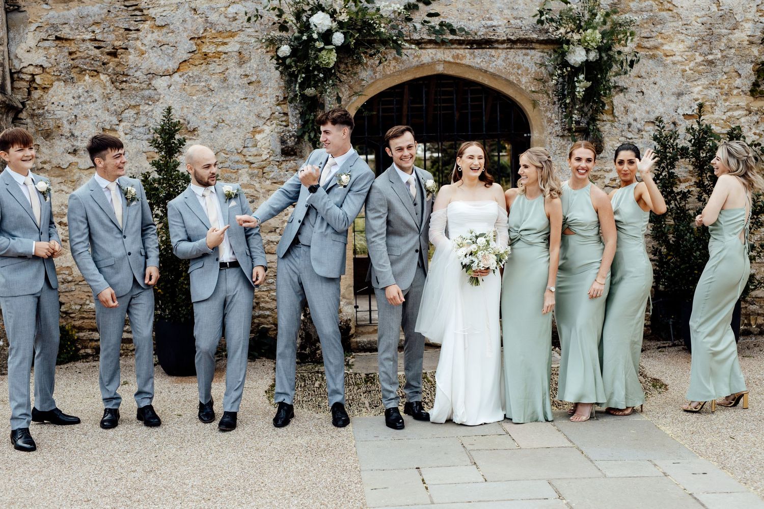 Wedding party in light blue and grey attire poses against stone wall with floral arrangements.