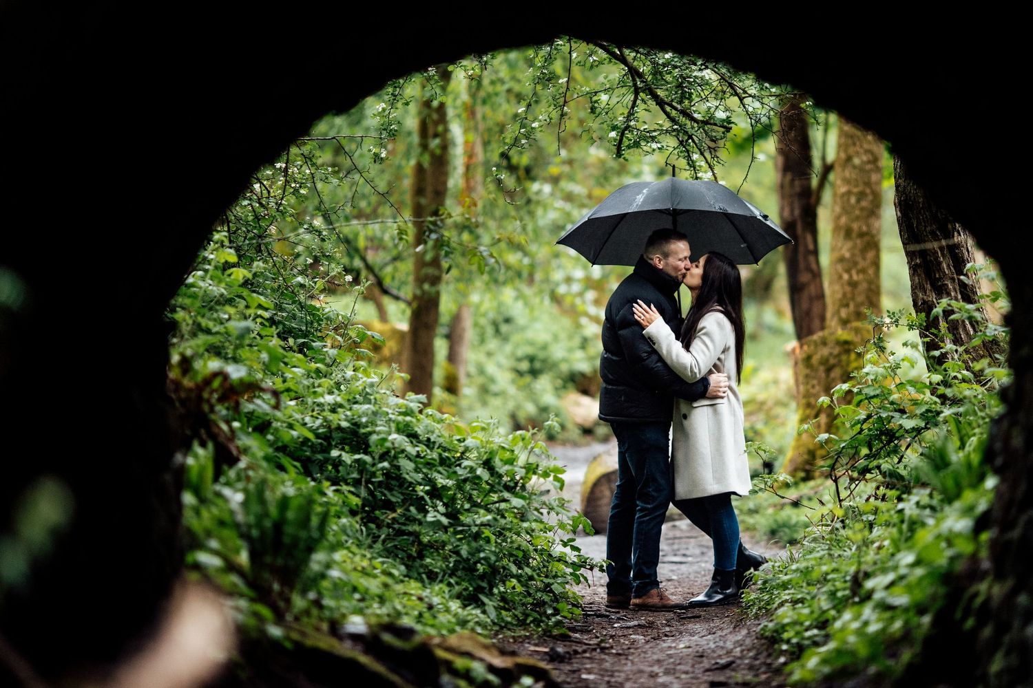 Two people share an intimate moment under a black umbrella in a misty forest setting.