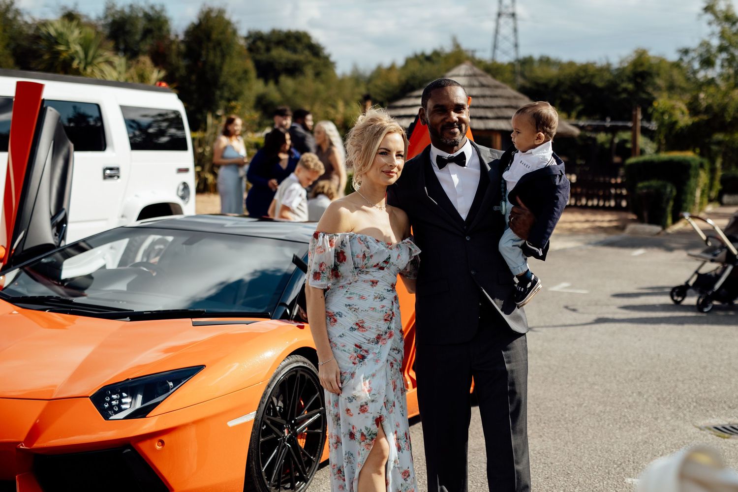 Orange sports car parked at wedding venue with wedding party gathered nearby.