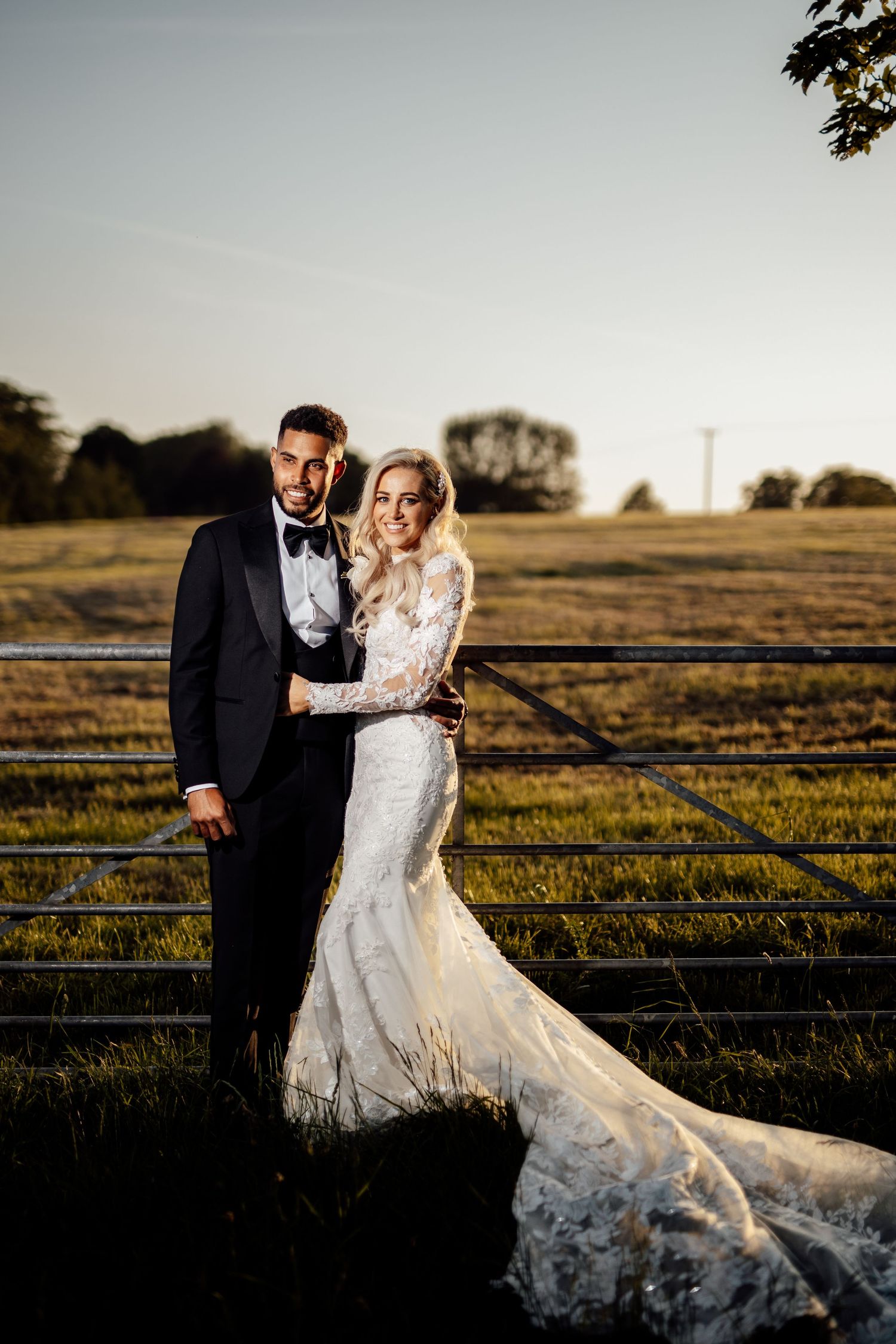 Couple poses in formal wedding attire against a sunset backdrop in a rural field.