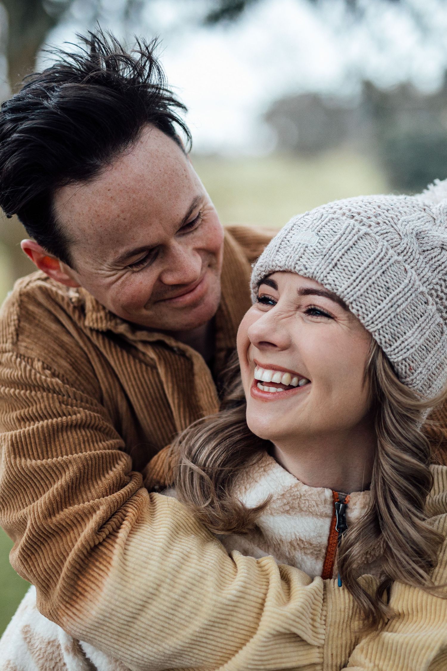 Playful couple laughing together while wearing winter attire in a natural outdoor setting.
