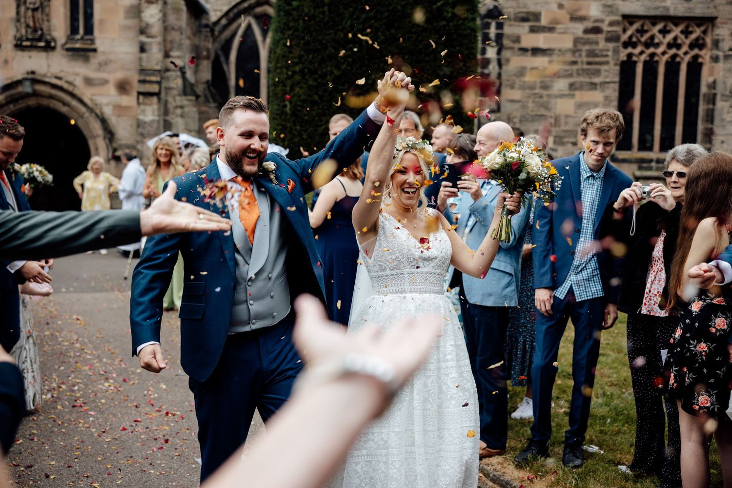 Wedding guests throw confetti as couple walks through stone courtyard.