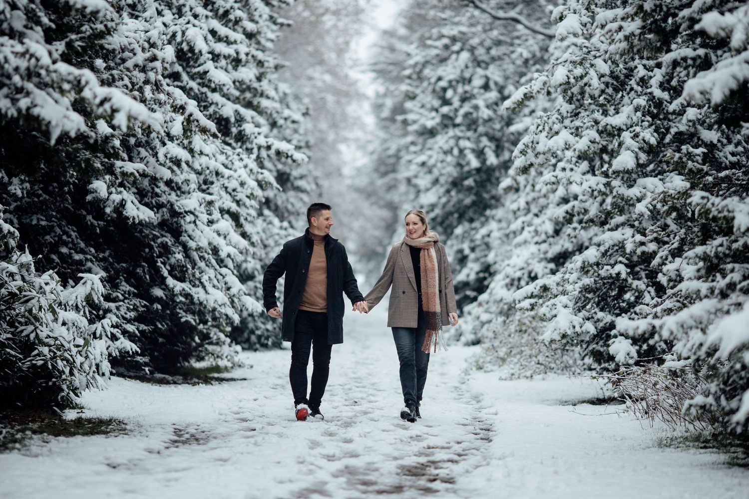 A couple walks hand in hand through a snow-covered evergreen forest path.