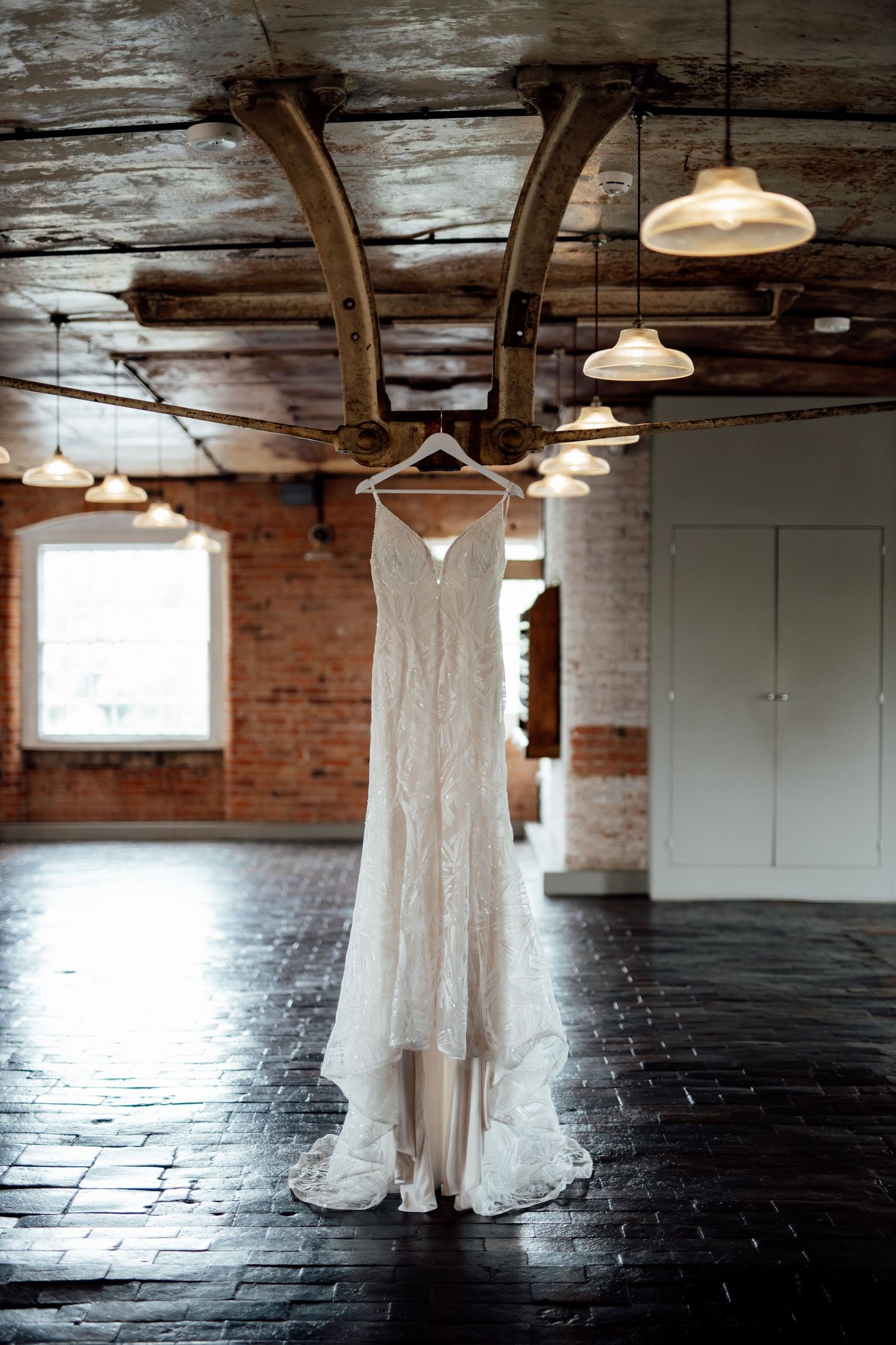 White lace wedding dress hanging in industrial brick venue with vintage pendant lights.