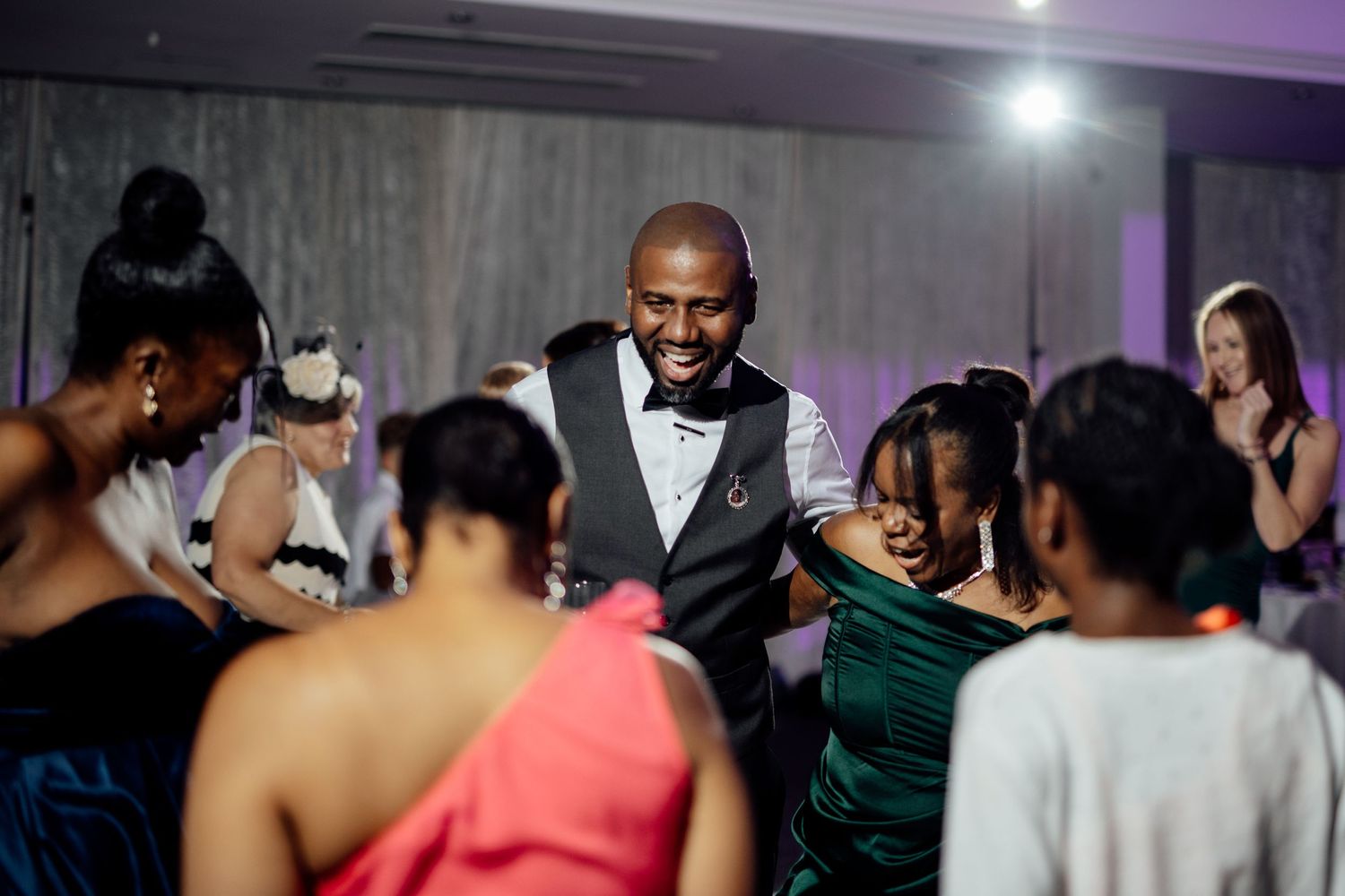 Wedding reception guests dance and celebrate under purple mood lighting.
