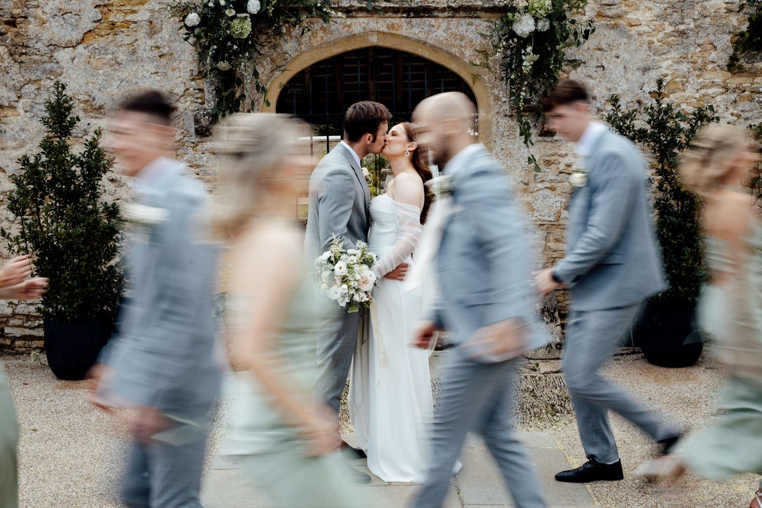 Creative motion blur shot of wedding party walking past stone wall with greenery.