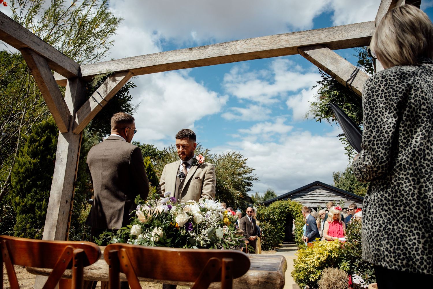 Groom stands at outdoor wedding altar under wooden arch, surrounded by floral arrangements and guests on a sunny day.