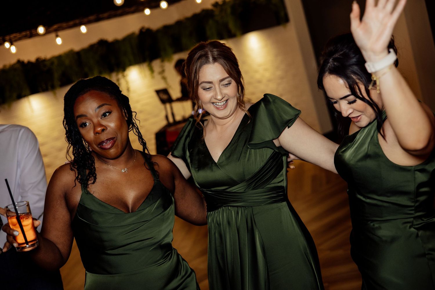 Three bridesmaids in emerald green satin dresses dancing joyfully at a wedding reception.
