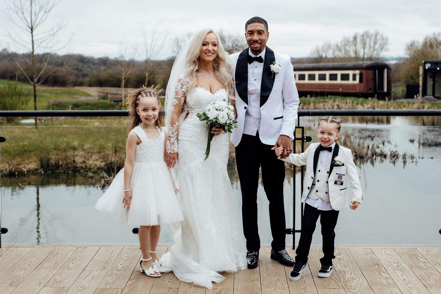 A bride and groom pose with a flower girl and ring bearer on a dock overlooking a serene pond.