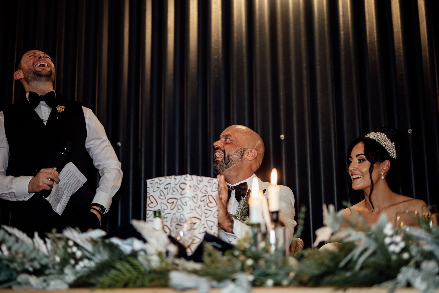 Wedding reception guests laughing and enjoying speeches at a dimly lit venue with modern decor.