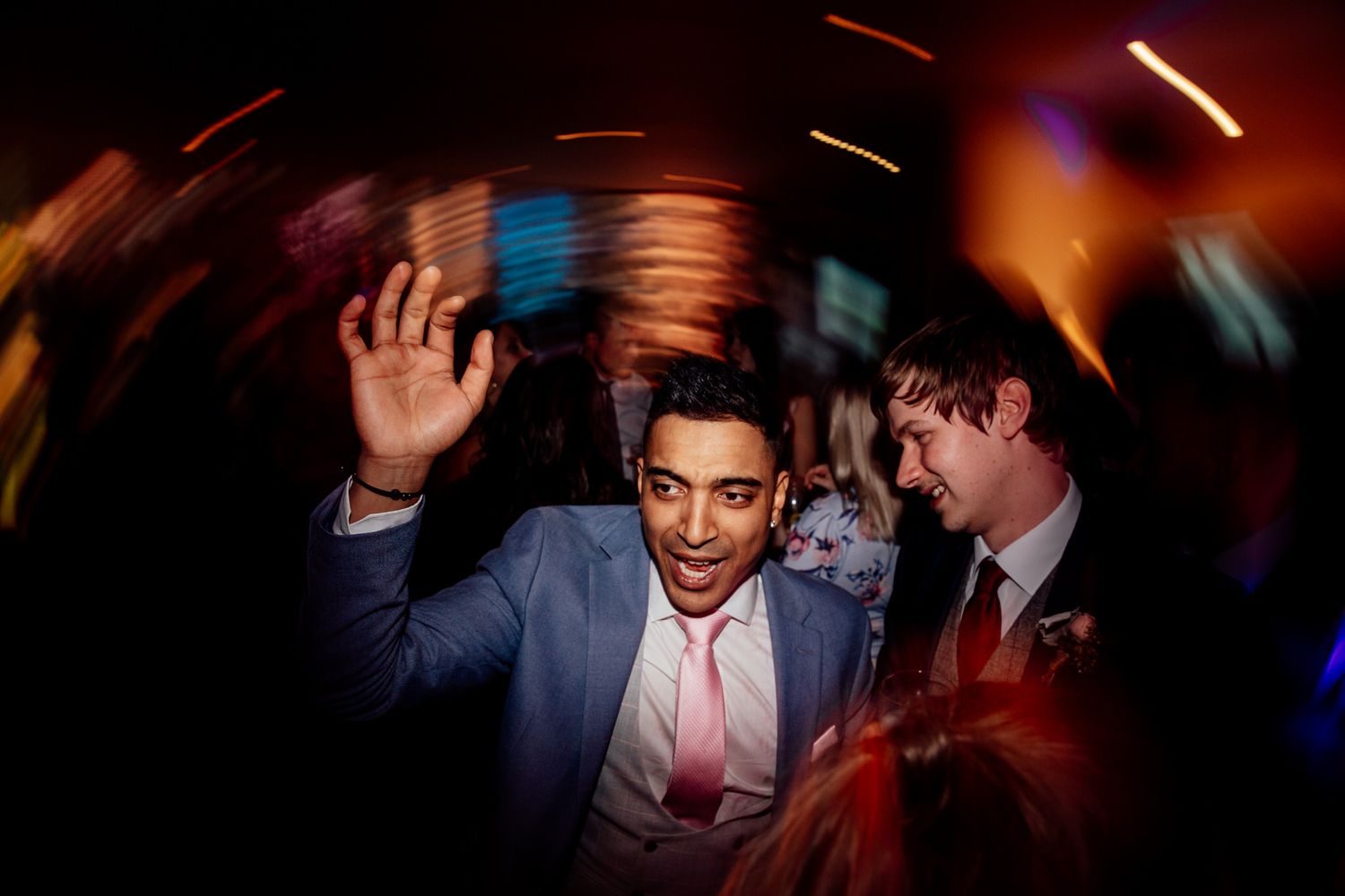 Wedding guests dancing and celebrating at an indoor reception with motion blur and warm lighting.