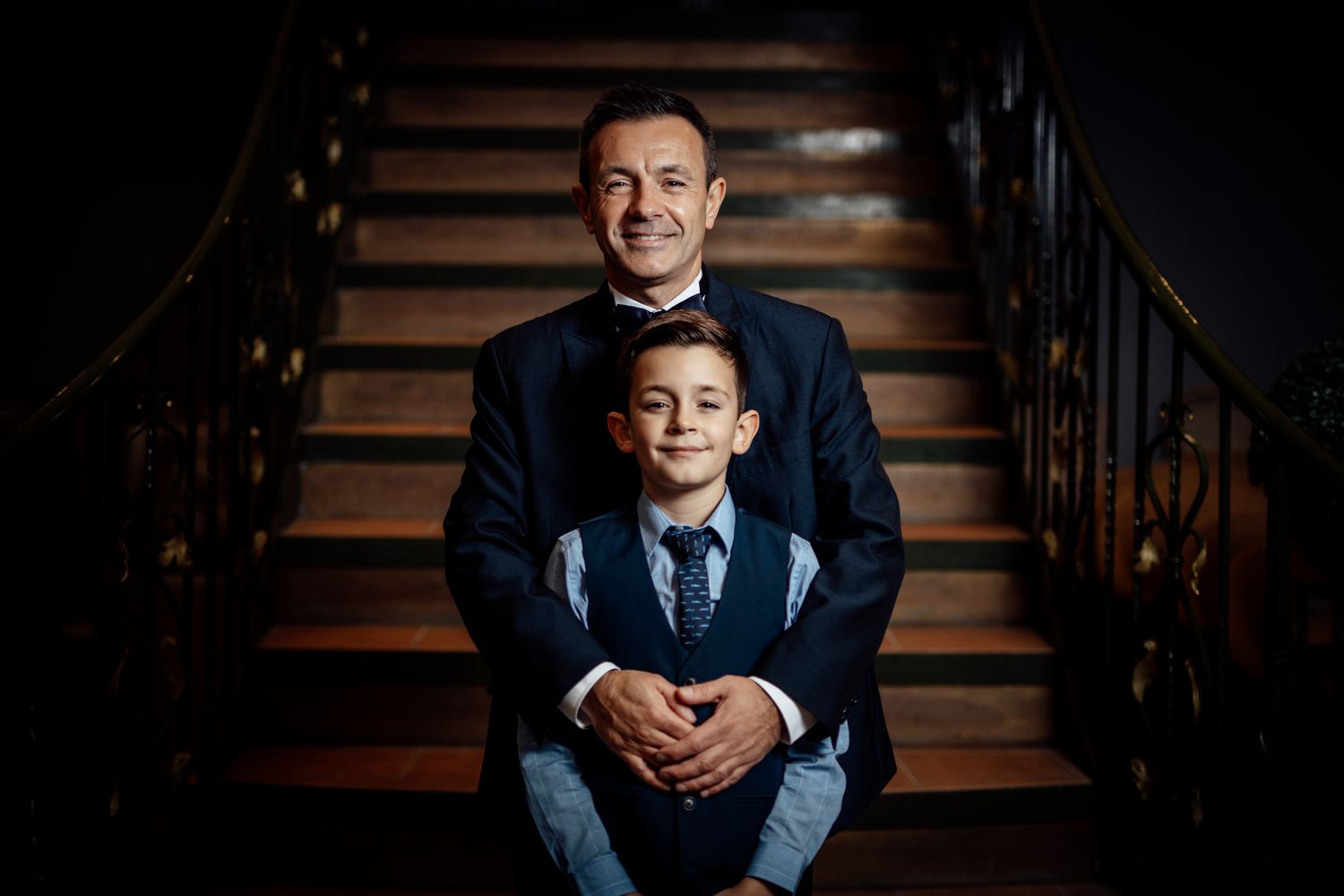 Formal portrait on wooden stairs with dramatic lighting and iron railing details.