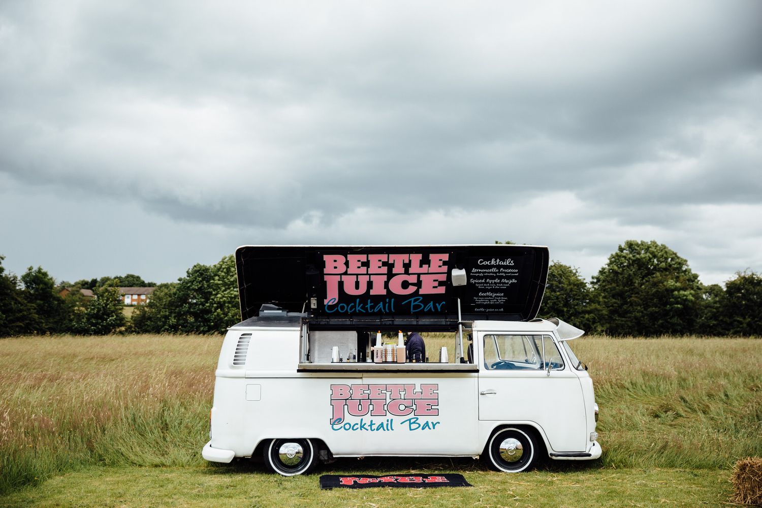 A vintage white VW van food truck with 'Beetle Juice' branding parked in a grassy field under cloudy skies.