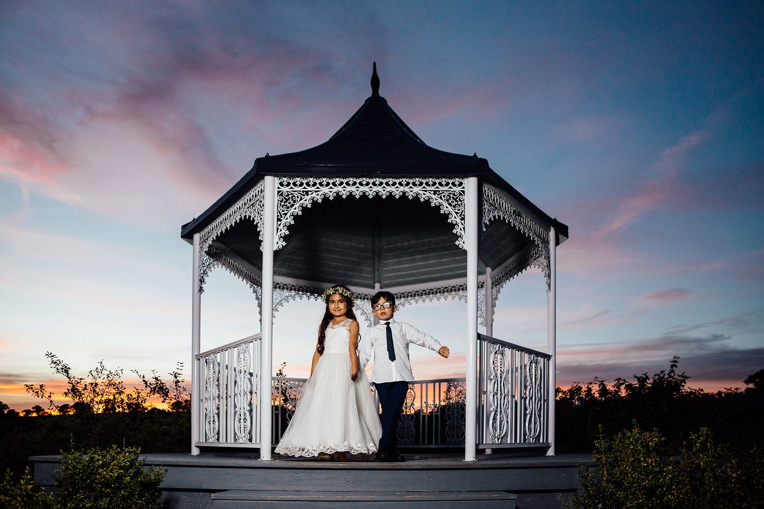 Two people in wedding attire stand in a white gazebo at sunset.