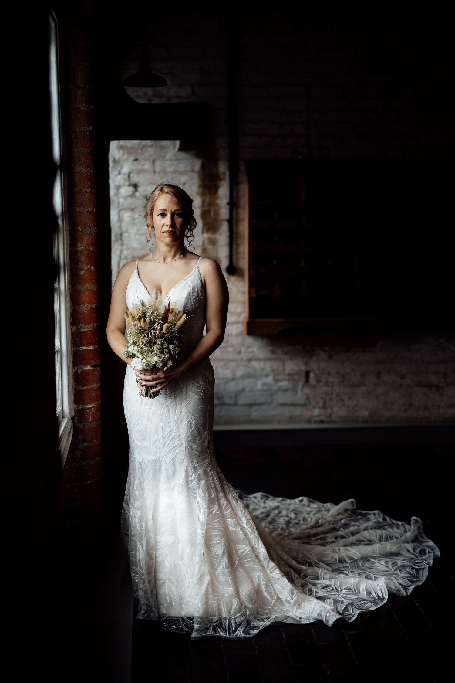 Elegant lace wedding dress with flowing train photographed in dramatic lighting against brick wall.