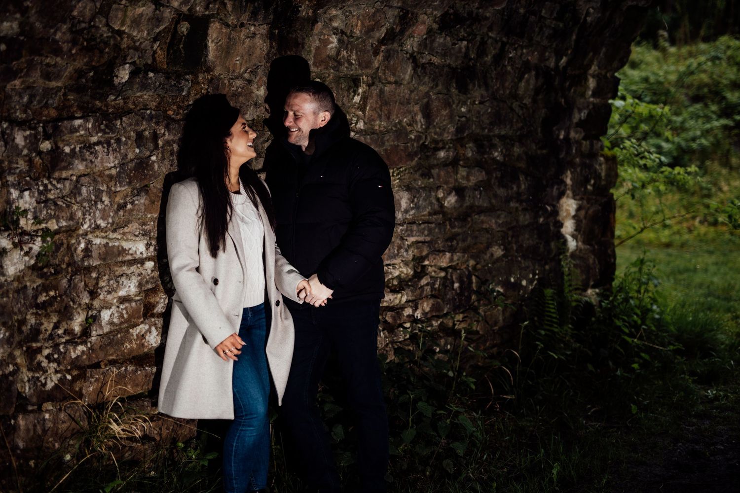 A romantic couple poses against a rustic stone wall in an outdoor setting.