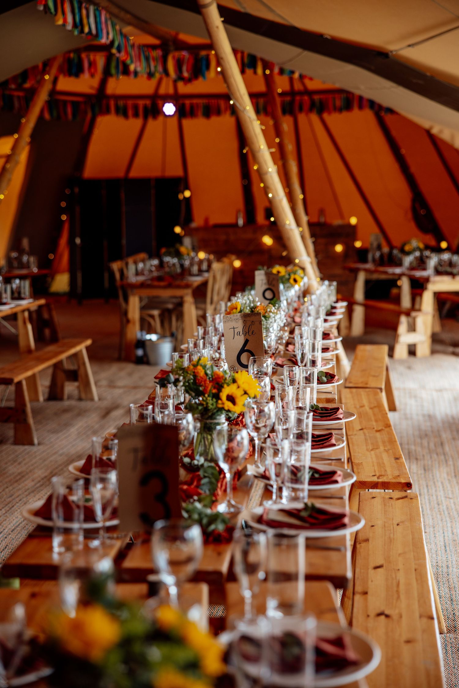 Rustic wedding reception tables set with glassware and floral centerpieces inside a tipi-style tent.