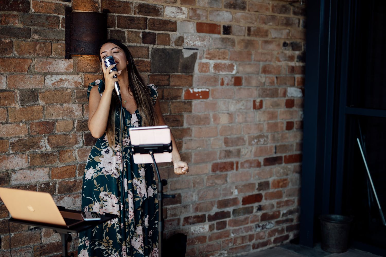 Woman in floral dress giving an emotional speech at brick-walled venue.