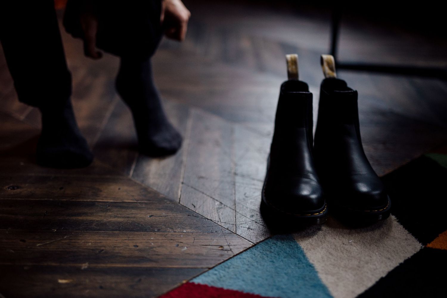 A pair of black leather chelsea boots on a wooden floor next to a geometric patterned rug.
